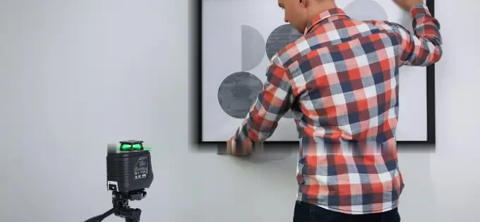 A framed picture being hung on a wall while a Firecore laser level on a tripod projects a horizontal green line along its bottom edge